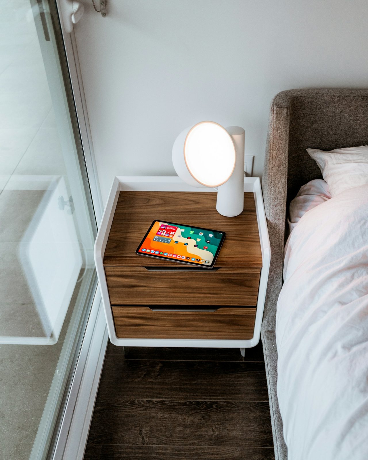 A compact bedside cabinet holding a round white lamp and a tablet next to white bedding.