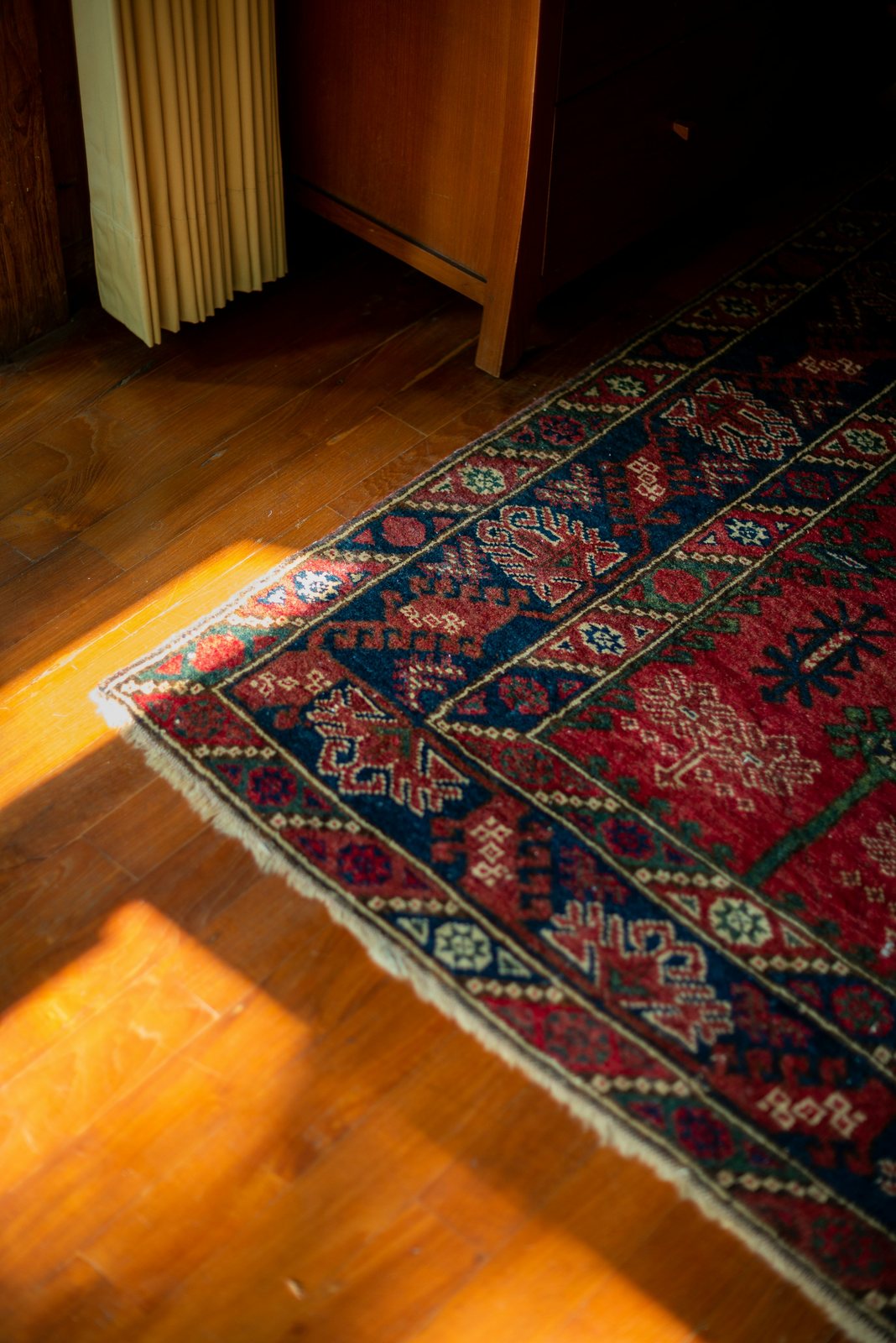 A patterned red and blue rug lies on a wooden floor beside a cabinet and radiator.
