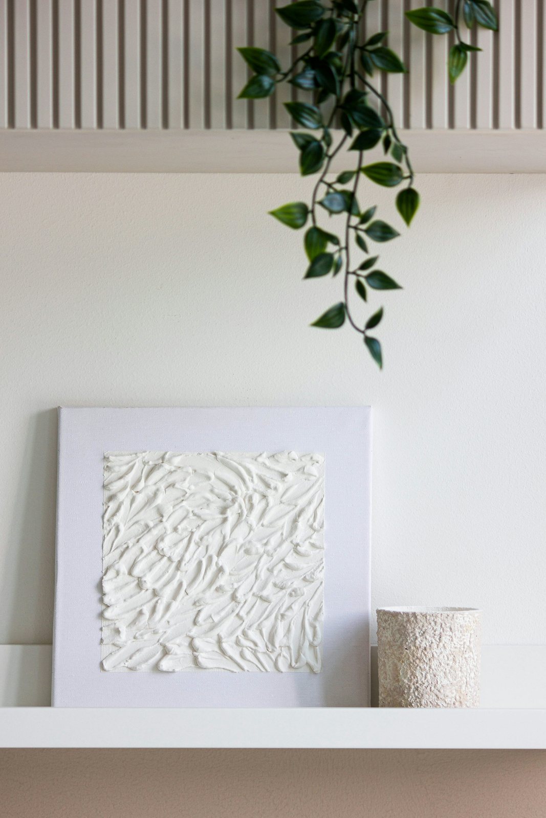 White textured artwork leans on a narrow shelf beside a small pot and a trailing plant.