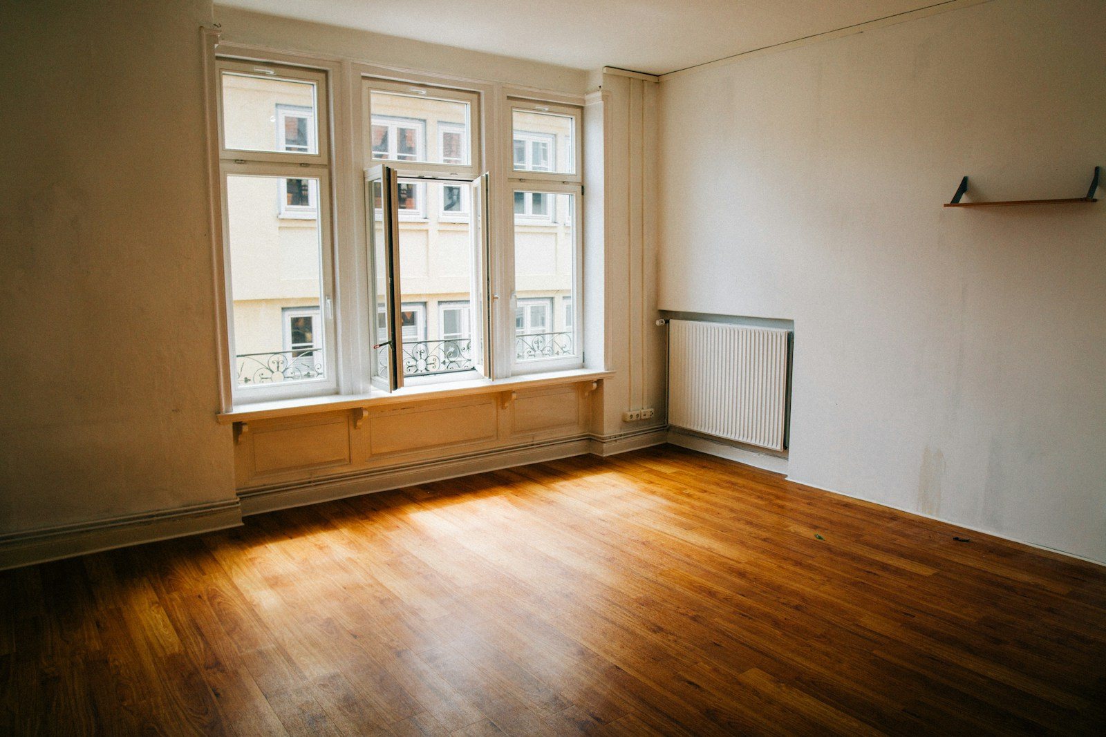 Sunlight across an empty wood floor below windows, with a radiator and a clear wall corner.