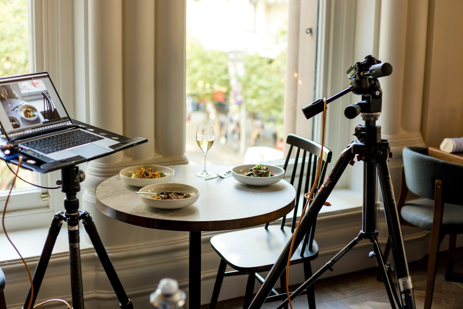 A laptop on a stand and a camera on a tripod beside a round table with bowls near a window.