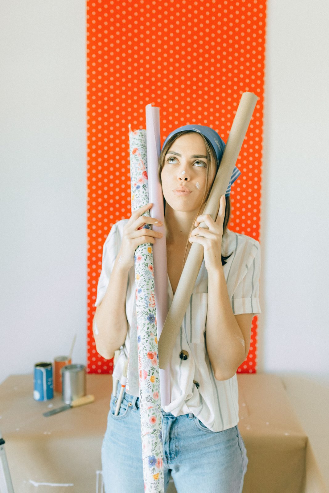 A woman holds several wallpaper rolls in front of a wall with an orange dotted wallpaper sample.