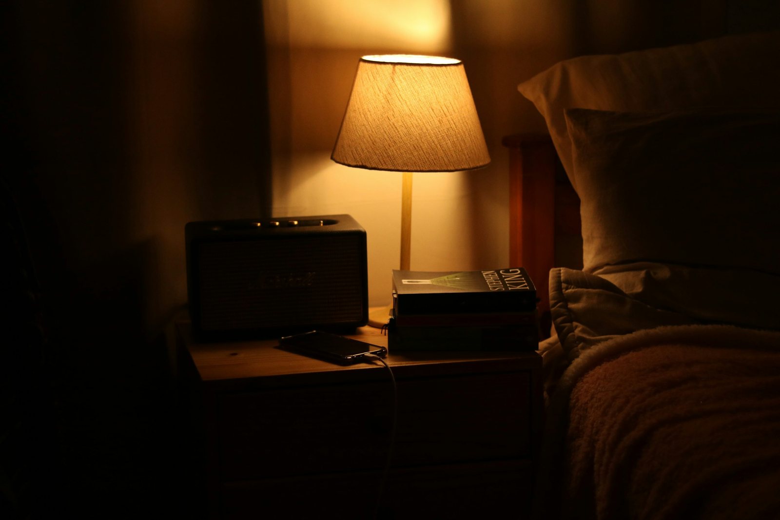 A warm table lamp illuminates books, a phone and a speaker beside a bed.