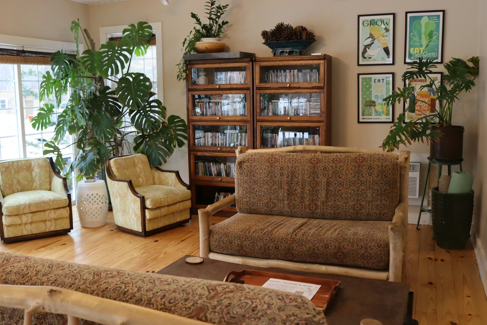 Patterned sofa and pale armchairs beside wooden book cabinets and leafy houseplants.
