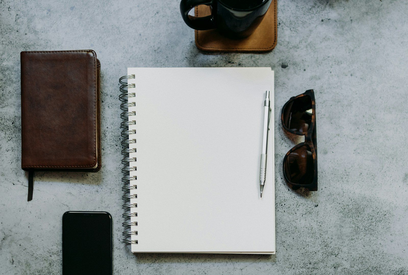 Blank spiral notebook and pen beside a phone, leather notebook, sunglasses and coffee on a gray table.