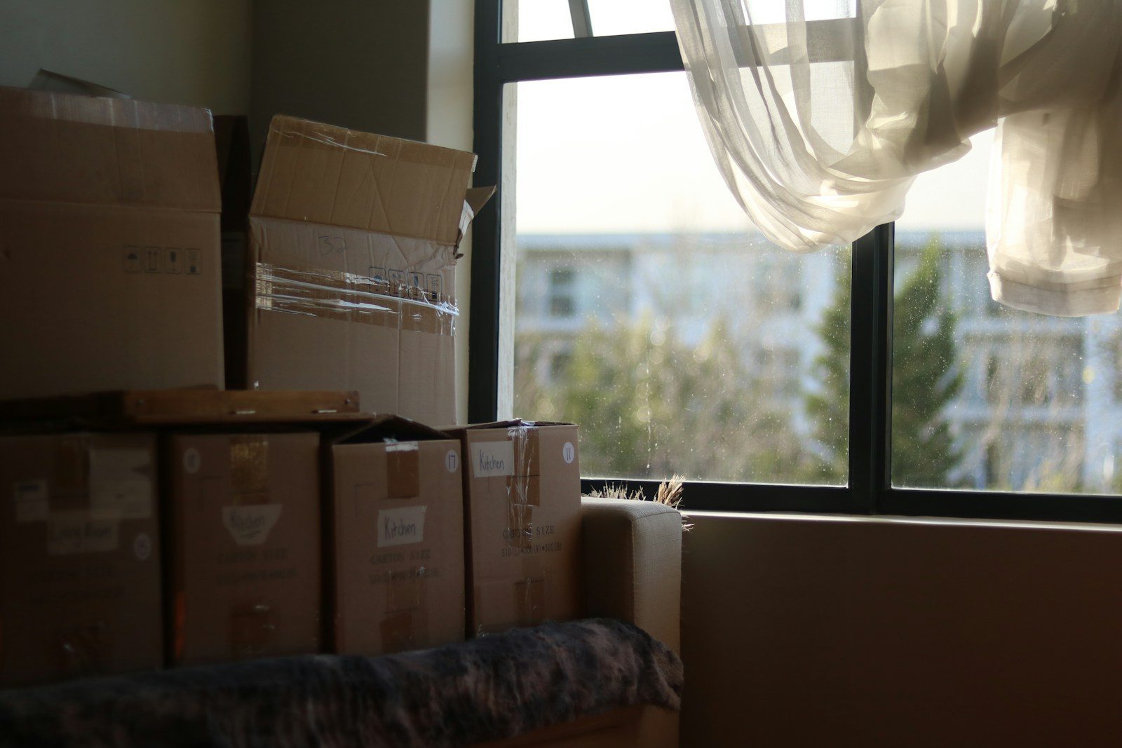 Used cardboard boxes stacked beside a sunlit window with a loose white curtain.