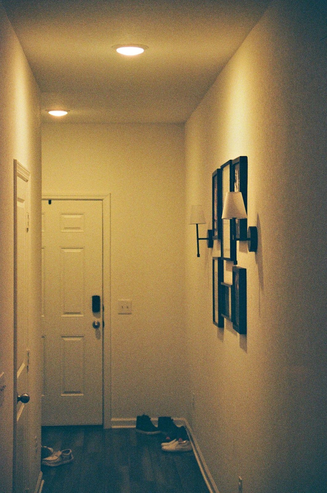 A narrow hallway with framed pictures, a white front door and shoes on the wood floor.
