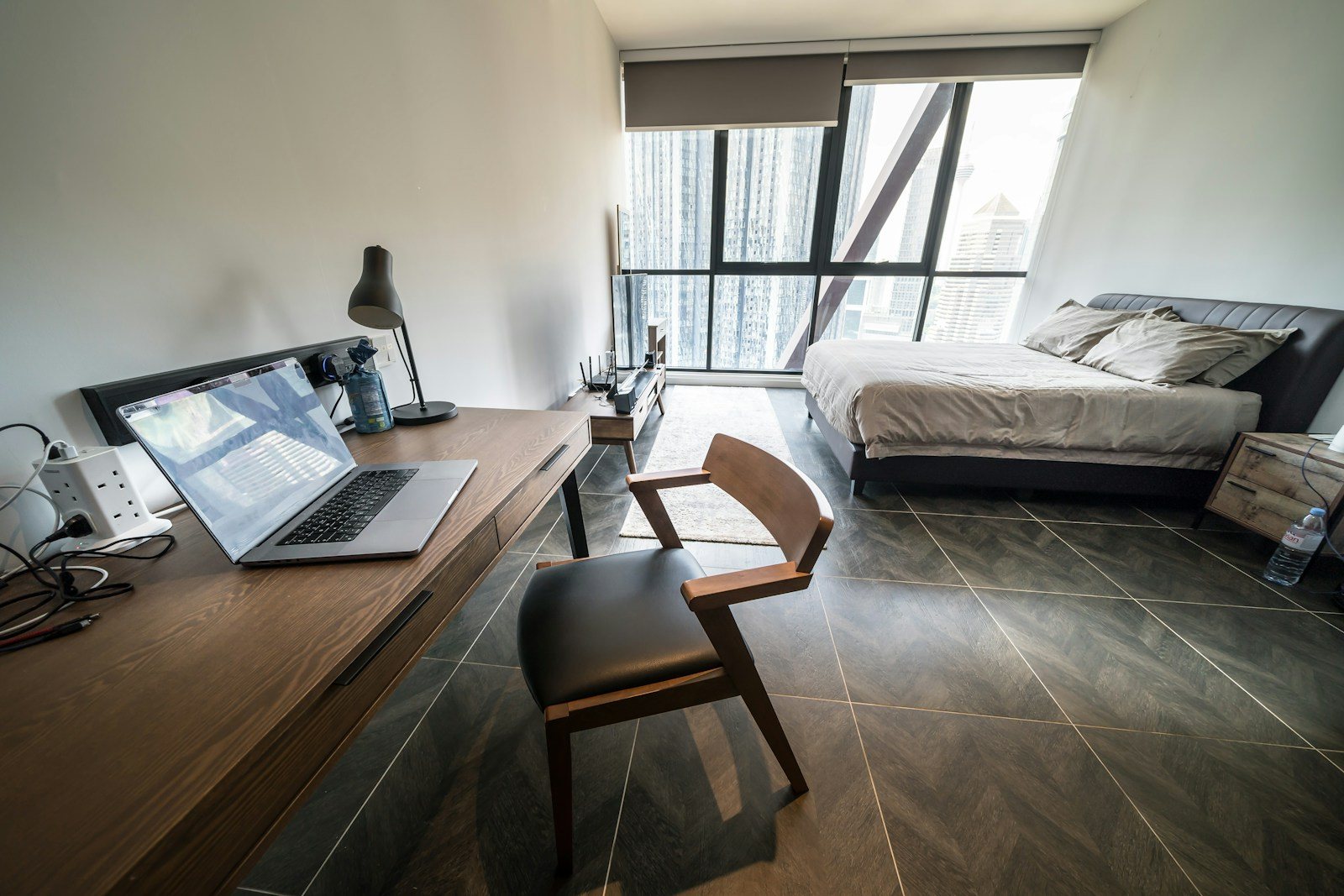 A desk with a laptop and chair beside a bed in a compact room with large windows.