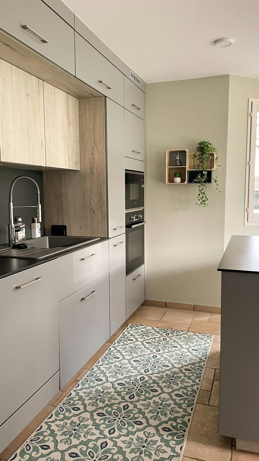 Compact kitchen with light cabinets, a wood backsplash, trailing plants and a green patterned runner.